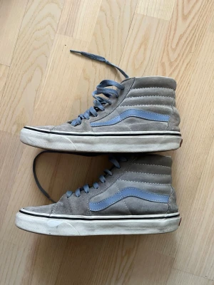Gråa Vans sneakers - Säljer ett par klassiska Vans Sk8-Hi sneakers i grå mocka med ljusblå sidestripe och matchande blå skosnören. Skorna har hög modell och vit sula med svart rand. Perfekta för en streetwear-look och passar till det mesta.