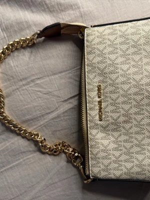 Beige kuvertväska Michael Kors - Stilren beige kuvertväska från Michael Kors med guldfärgad kedjerem och dragkedja. Väskan har präglat mönster, logga framtill och flera fack inuti. Perfekt för att bära det viktigaste med snygg detalj.