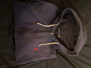 Mörkblå hoodie från Polo Ralph Lauren, Storlek M - Mörkblå hoodie med dragkedja från Polo Ralph Lauren. Klassisk design med vit dragsko i huvan, två fickor framtill och röd broderad logga på bröstet. Mjuk bomullskänsla, perfekt för en avslappnad stil. Väldigt bra sick. 