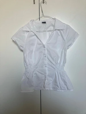 Vit blus från Gina Tricot - Fräsch vit blus från Gina Tricot med korta ärmar, krage och knäppning framtill. Blusen har smock i midjan för en snygg passform. Perfekt till jeans eller kjol för en clean look. Andvänd en gång.