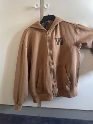 Brun hoodie med dragkedja och W-tryck - Snygg brun hoodie med dragkedja och huva. Framtill finns ett broderat W på bröstet och två stora fickor. Hoodien har ribbade muddar vid ärmslut och nederkant. Perfekt för en chill och avslappnad stil.