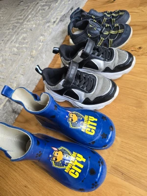 Blå Paw Patrol gummistövlar barn - Säljer ett par blå gummistövlar för barn med Paw Patrol-motiv och texten 'Save the City'. Stövlarna har svarta tassavtryck och är tillverkade i vattentätt material med elastisk resår vid skaftet för enkel påtagning.