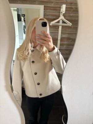 Beige ulljacka med stora knappar - Snygg beige jacka i ullmix med stora bruna knappar och klassisk krage. Jackan har raka ärmar, två sidofickor och justerbara band vid ärmsluten. Perfekt för dig som vill ha en stilren och enkel look. Använd endast en gång och är perfekt för vår och höst. 🤍🤎