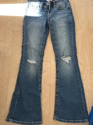 Blå bootcut jeans med slitningar - Säljer ett par blå bootcut jeans med slitningar på knäna och snygga detaljer vid fickorna. Jeansen har klassisk femficksdesign, dragkedja och knapp i midjan samt kontrastsömmar. Perfekta för en avslappnad och trendig look.storlel 164