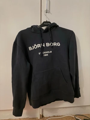 Svart hoodie från Björn Borg - Cool svart hoodie från Björn Borg med vit text på bröstet som säger 'BJÖRN BORG STOCKHOLM 1956'. Hoodien har en stor ficka framtill, huva med dragsko och ribbade muddar. Perfekt för en avslappnad och sportig look.