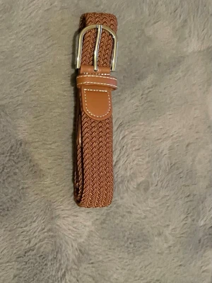 Brunt flätat bälte med skinn-detaljer - Snyggt brunt flätat bälte med detaljer i skinn och silverfärgat spänne. Perfekt accessoar för att lyfta din outfit. Bältet har en klassisk design med vita sömmar på skinnpartierna och passar till både jeans och byxor.obs går att upphämtas i södra Stockholm 