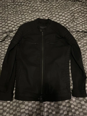 Svart denim jacka med dragkedjor - Snygg svart denim jacka med raka linjer och coola detaljer som dragkedjor på bröstet och quiltade partier på axlarna. Jackan har ståkrage och stängs med dragkedja framtill. Perfekt för en stilren och edgy look. Aldrig använd.