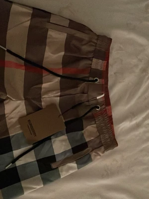 Rutig shorts från Burberry med snörning - Säljer ett par klassiska Burberry shorts i den ikoniska rutiga designen med beige, bruna, svarta och röda toner. Shortsen har elastisk midja med svart snörning och är tillverkade i ett lätt och bekvämt material. Perfekta för sommaren!