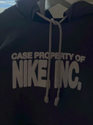 Grå hoodie från Nike med tryck - Mörkgrå hoodie från Nike med vit text 'CASE PROPERTY OF NIKE, INC.' på bröstet. Klassisk känguruficka framtill och justerbar huva med vita snören. Perfekt för en avslappnad streetwear-look.
