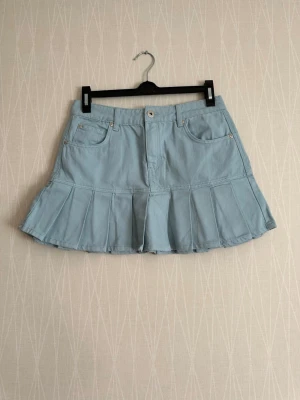 Ljusblå plisserad skort i denim - Trendig ljusblå skort i denim med plisserad nederdel och klassiska jeansdetaljer som fickor fram och bak samt hällor för skärp. Perfekt för en cool och avslappnad stil. Stängs med knapp och dragkedja framtill.