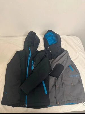 Svart och grå vindjacka från Funk - Två snygga vindjackor från Funk, en svart med blå detaljer och en grå med svarta och blå detaljer. Båda har huva, dragkedjor i kontrastfärg och flera fickor. Perfekta för blåsiga dagar och outdoor-aktiviteter.