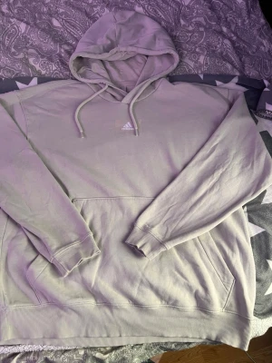 Ljusgrå hoodie från Adidas - Snygg ljusgrå hoodie från Adidas med klassisk logga på bröstet. Hoodien har en stor känguruficka framtill, justerbar dragsko i huvan och ribbade muddar vid ärmar och nederkant. Tillverkad i mjukt bomullsmaterial för skön känsla.