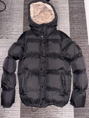 Alessandro Zavetti Delucci Puffer 2 in 1 jacka - Jetblack (svart) dunjacka från Alessandro med avtagbara ärmar som gör att den kan användas som väst. Jackan har en beige innerfoder, stor huva och dragkedja med knappar. Två stora fickor med dragkedja framtill. Perfekt för kalla dagar.  Nypris: 1495kr