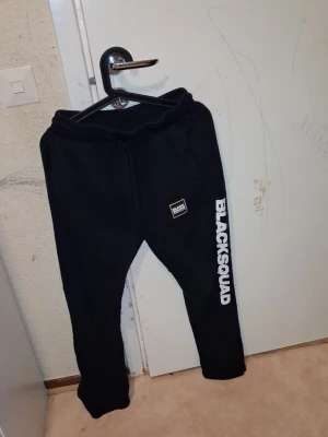 Svarta mjukisbyxor BLACKSQUAD (Man) - Svarta mjukisbyxor från BLACKSQUAD med vit logga på ena benet och liten patch framtill. Byxorna har snörning i midjan och är i en loose passform, perfekta för chill och träning. Materialet är mjukt och bekvämt, troligen bomull eller polyester.