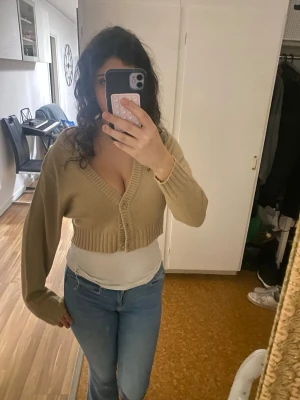 Beige cropped kofta med knappar - Säljer en knappast använd beige cropped kofta med djup v-ringning och breda ribbade muddar. Koftan har tre knappar framtill och långa ärmar. Perfekt att slänga över ett linne eller t-shirt för en chill look.