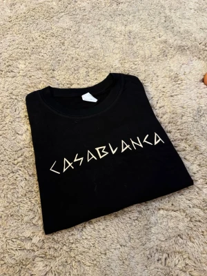  Casablanca t-shirt  - Snygg svart t-shirt från Casablanca med vit grafisk logga på bröstet. Klassisk rund hals och korta ärmar. Tillverkad i mjuk bomull som känns skön mot huden. Perfekt för dig som gillar stilrena och trendiga plagg.