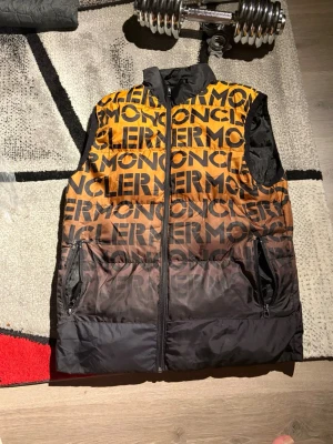 Moncler dunväst med logotypmönster (S-M) - Snygg dunväst från Moncler med fet logotyp över hela västen. Färgen går i en cool gradient från orange upptill till svart nedtill. Västen har dragkedja framtill och två fickor med dragkedja. Perfekt för dig som vill sticka ut med streetstyle.
