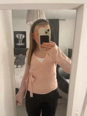 Ljusrosa omlottstickad tröja  - Supersöt ljusrosa stickad omlottopp med knytband i sidan och långa ärmar. Tröjan har en mjuk och fluffig känsla, perfekt för lager på lager. Passar till både jeans och kjol för en trendig look. den är från lager 157 i storlek xs.