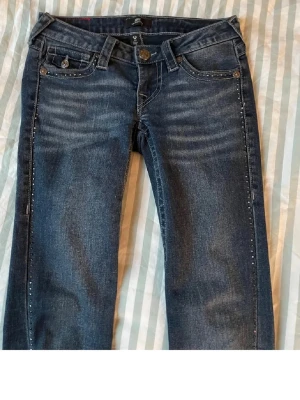 Mörkblå jeans med nitar - Snygga mörkblå jeans med raka ben och dekorativa nitar längs fickor och sidor. Jeansen har klassisk femficksdesign och markerade sömmar på bakfickan. Tillverkade i denim med lätt tvättade detaljer för en cool look.