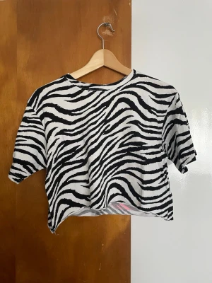 Zebramönstrad croppad t-shirt - Cool croppad t-shirt med svartvitt zebramönster och stora neonrosa tryck på ryggen med texten 'JACOBS' och siffran 63. Perfekt statement-plagg för dig som vill sticka ut. T-shirten har korta ärmar och avslappnad passform.