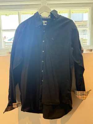 Svart skjorta från Burberry - Säljer en ikonisk svart skjorta från Burberry i storlek L. Den har det Ikoniskt rutigt mönster på insidan av manschetterna och märket broderat på bröstet. Nypris 4500. Säljs endast för 699