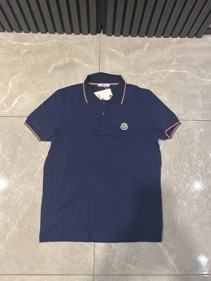 Marinblå polotopp från Moncler - Säljer en marinblå polotopp från Moncler med klassisk krage och tre knappar framtill. Toppens krage och ärmslut har snygga röda och vita detaljer. Moncler-loggan är broderad på bröstet. Perfekt för dig som gillar stilrena och exklusiva plagg.