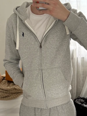 Grå zip-hoodie från Polo Ralph Lauren - Snygg grå zip-hoodie från Polo Ralph Lauren med klassisk huva och vita snören. Hoodien har två fickor framtill, ribbade muddar och det ikoniska Polo-logot broderat på bröstet. Perfekt för en chill och avslappnad look.