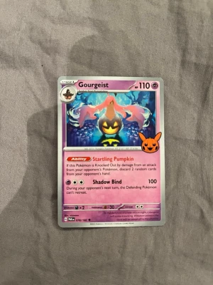 Pokémonkort Gourgeist - Säljer ett Pokémonkort med motivet Gourgeist. Kortet har lila och rosa färgtoner, med en pumpaliknande figur i centrum och ett Pikachu-huvud på framsidan. Text och symboler i flera färger. Perfekt för samlare eller Pokémonfans.