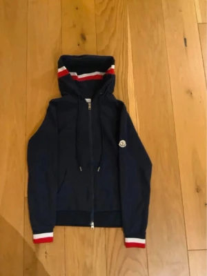 Marinblå Moncler hoodie med dragkedja - Snygg marinblå hoodie från Moncler med dragkedja framtill och huva. Röd och vit rand på huvan och ärmsluten ger en sportig vibe. Klassisk Moncler-logga på ärmen. Perfekt för dig som gillar streetwear och exklusiva märken.