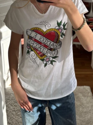 Zadig Tshirt! Vit t-shirt med hjärtmotiv - Vit t-shirt med färgglatt hjärtmotiv och blommor på bröstet. Trycket har röda, gula och gröna detaljer och text i banderollstil. T-shirten har korta ärmar och en lätt v-ringad hals. Perfekt för en cool och avslappnad stil.
