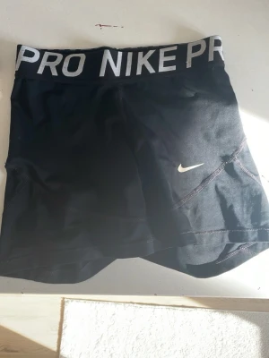Svarta Nike Pro träningsshorts - Svarta Nike Pro shorts med bred resår i midjan där det står 'NIKE PRO' i vitt. Tight passform och korta ben, perfekta för träning. Materialet är stretchigt och andas, med en liten vit Nike-logga på benet.