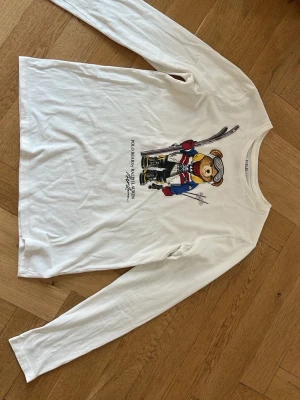 Vit långärmad topp Polo Bear - Vit långärmad topp från Ralph Lauren med ikoniskt Polo Bear-tryck framtill. Toppen har rund halsringning och är gjord i mjuk bomull, perfekt för en chill och sportig look.