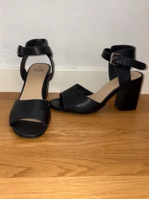 Svarta chunky klackskor  - Snygga svarta pumps från Vox med öppen tå, bred klack och justerbart spänne runt ankeln. Skorna har en stilren design i konstläder och passar perfekt till dig som gillar chunky klackar och enkel stil.