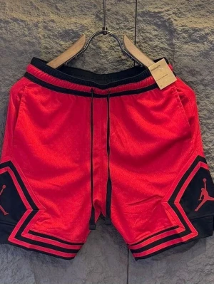 Röda Jordan shorts med svarta detaljer - Snygga röda shorts från Jordan med svarta detaljer och logga på båda benen. Shortsen har snörning i midjan och är perfekta för en sportig look. Tillverkade i ett lätt och ventilerande material som passar bra till sommaren.