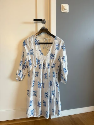 Vit blommig klänning med knyt - Söt vit klänning med blått blommönster och v-ringning. Klänningen har trekvartsärm och knytband i nacken. 