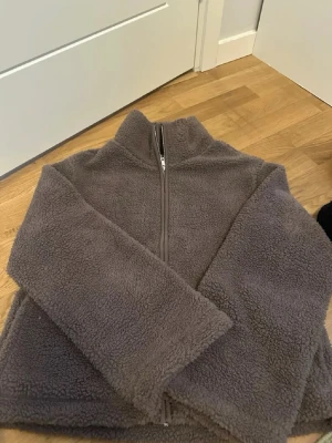 Beige  Pilejacka med dragkedja stl M  - Mysig mörkgrå teddyjacka med hög krage och hel dragkedja framtill. Jackan är tillverkad i fluffigt teddy-material och har en avslappnad passform. Perfekt för kalla dagar när du vill vara både varm och stilren. Helt ny med etiketterna kvar :) 