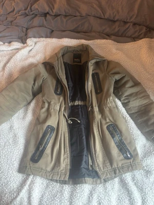 Beige parkas från Outfitters Nation - Stilren beige parkas från Outfitters Nation med svarta detaljer och dragkedjor på fickorna. Jackan har dragsko i midjan för snygg passform och svart innerfoder. Perfekt för kyliga dagar och enkel att matcha med olika outfits.
