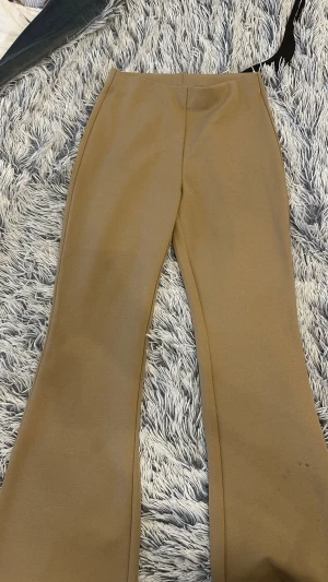 Beige utsvängda kostymbyxor - Snygga beige kostymbyxor med utsvängda ben och hög midja. Byxorna har en stilren design utan synliga fickor eller detaljer och är gjorda i ett mjukt, stretchigt material som ger en skön passform. Perfekta för en clean och trendig look.