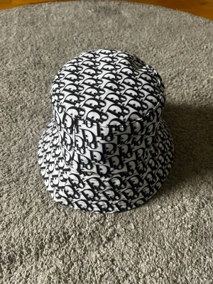 Svartvit bucket hat från Dior - Säljer en svartvit bucket hat från Dior med det klassiska Dior-monogrammet över hela hatten. Hatten har en rund form och bred skärm, tillverkad i ett mjukt tyg som känns bekvämt på huvudet. Perfekt accessoar för att lyfta din outfit.