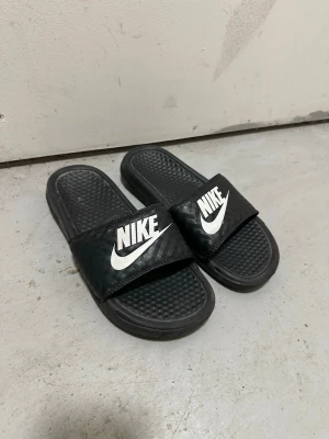 Svarta Nike badtofflor - Svarta sandaler från Nike med bred rem över foten och vit logga. Klassisk slip-on modell med räfflad sula för extra grepp. Tillverkade i syntetmaterial och perfekt för chill dagar eller efter träningen.