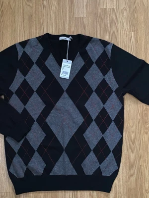 Argylemönstrad tröja  - Snygg stickad tröja från Dressmann med klassiskt argylemönster i svart, grått och tunna röda linjer. Tunnare tröja. Tröjan har V-ringning hel svart baksida. Helt ny med lappar och från ett djur och rökfritt hem! 