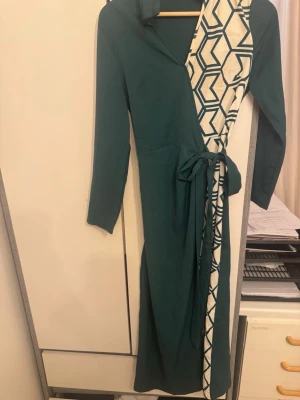 Grön omlottklänning med mönster - Snygg lång grön omlottklänning med långa ärmar och knyt i midjan. Ena sidan har ett vitt geometriskt mönster i grönt som ger en cool kontrast. Perfekt för dig som gillar unika detaljer och stilren design.