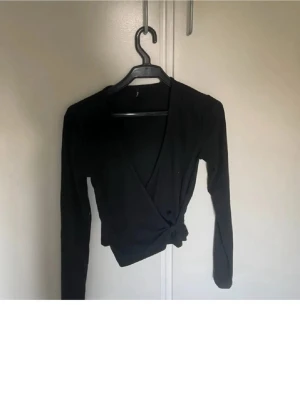 Svart omlottopp med knyt - Snygg svart omlottopp med lång ärm och knyt i sidan. Toppen har en v-ringad design vilket ger en trendig look. Perfekt att matcha med  jeans eller kjol.