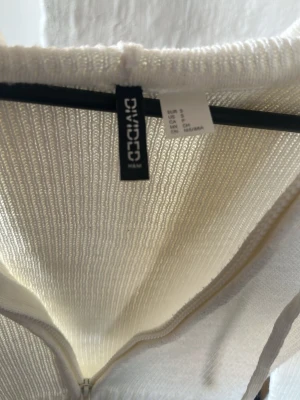 Vit stickad hoodie från Divided - Vit stickad  från Divided med dragkedja framtill och ribbstickade detaljer nedtill. Tröjan har huva med snören och är perfekt för en avslappnad stil. Materialet är mjukt och lätt, vilket gör den skön att bära.