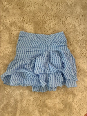 Minikjol - Supersöt blå kjol med volanger och tunna vertikala ränder. Kjolen har en luftig och lätt känsla, perfekt för varmare dagar. Tillverkad i ett tunt, transparent tyg med flera lager för extra volym och rörelse.