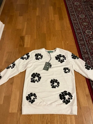 Vit sweatshirt från Denim Tears - Vit sweatshirt från Denim Tears med svarta blommönster över hela tröjan. Rund halsringning och långa ärmar. Tillverkad i mjukt bomullsmaterial och har en avslappnad passform. Perfekt för dig som gillar streetwear och unika prints.