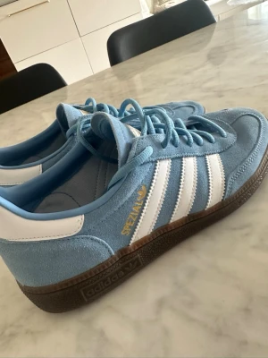 Adidas Spezial blå sneakers - Snygga Adidas Spezial sneakers i ljusblå mocka med vita detaljer och klassiska tre ränder på sidan. Skon har brun gummisula och guldfärgad text.