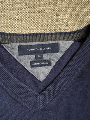 Mörkblå stickad tröja Tommy Hilfiger - Mörkblå stickad tröja från Tommy Hilfiger med klassisk V-ringning och diskret broderad logga på bröstet. Tröjan har ribbade muddar vid hals, ärmslut och nederkant. Perfekt för dig som gillar stilrena och tidlösa plagg.