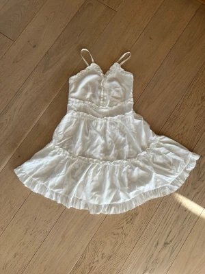 Design by Amor volangklänning S - Supersöt vit kortklänning med smala axelband och volangdetaljer. Klänningen har omlottkänsla upptill, knappar framtill och en luftig kjol med flera lager volanger. Perfekt för sommarens alla tillfällen!