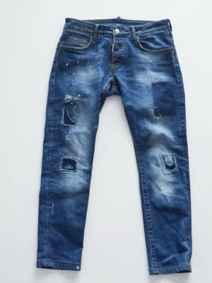 DSQUARED2 Jeans - Säljer ett par blå jeans med slitningar, patchdetaljer och färgstänk för en cool och trendig look. Jeansen har klassisk femficksmodell och raka ben. Perfekta för dig som gillar streetstyle och vill sticka ut lite extra.